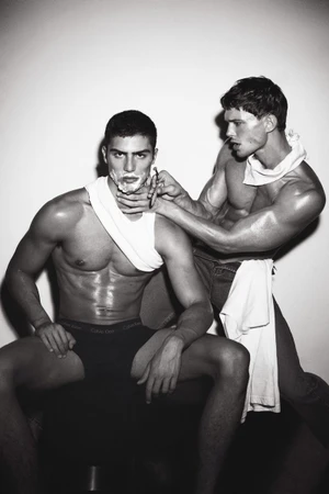 Two shirtless men in underwear, one sitting with shaving cream on his face while the other holds a razor, against a white background.