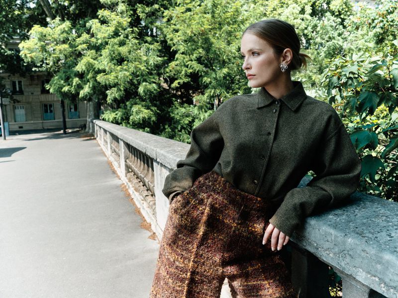 Woman in tweed outfit poses on bridge outdoors, wearing collared shirt and statement earrings, with green foliage in soft natural light.