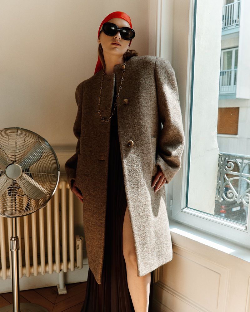 A woman wearing a coat, sunglasses, and a headscarf stands indoors in front of a window with a vintage fan to the left.