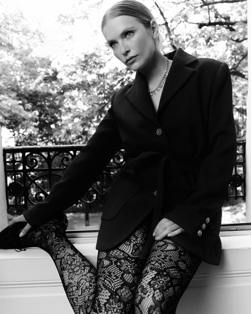 A blonde woman wearing a black blazer and patterned tights poses near a window with green foliage in the background, creating a high-fashion, monochromatic look.