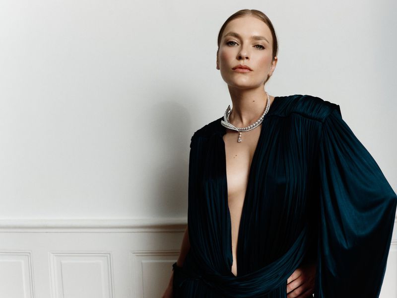 Elegant woman in a dark teal gown and diamond necklace poses against a white wall, creating a sophisticated fashion image.