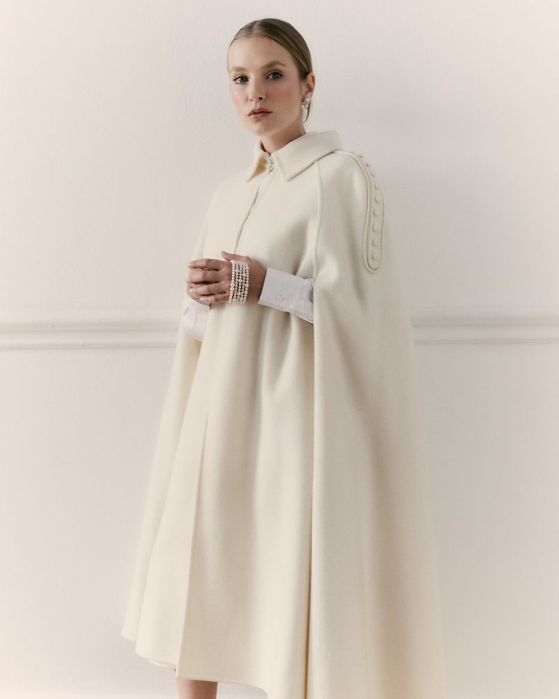 Elegant woman wearing a cream-colored cape stands in front of a white wall in a fashion studio. She has a neutral facial expression.