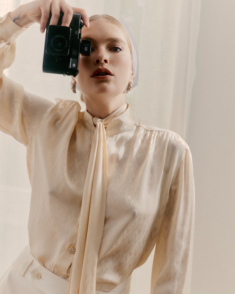 Blonde woman in a cream blouse holding a vintage camera to her eye, posing in front of a neutral wall in a portrait-style shot.