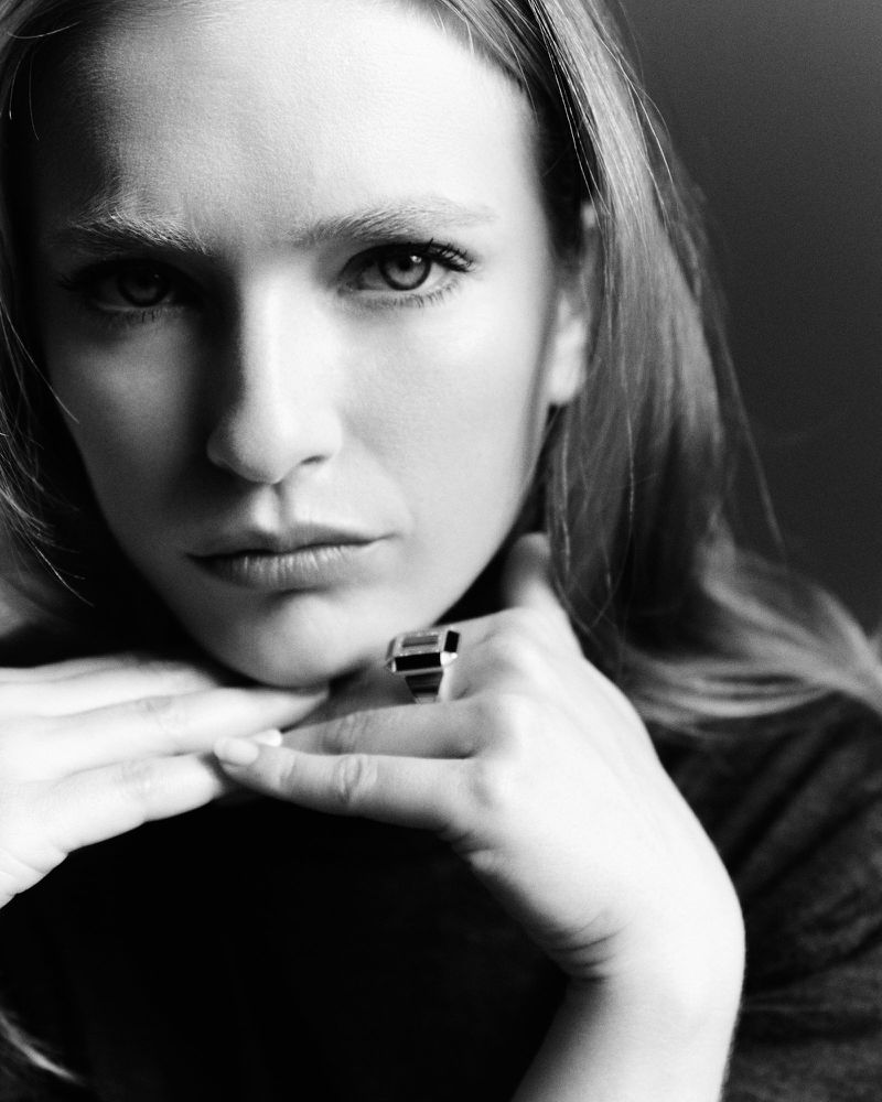 Monochrome portrait of an adult woman with light hair, fair skin, wearing a large ring, resting her chin on her interlaced fingers.