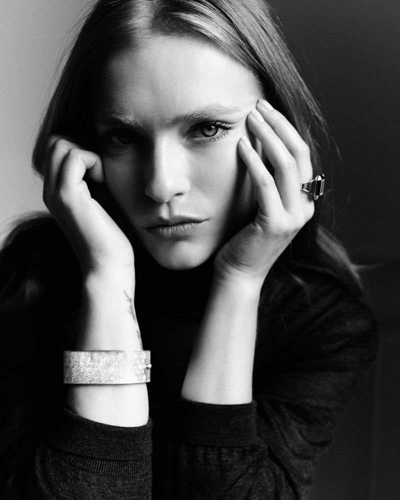 Monochrome portrait of a female model with hands on her face, showcasing jewelry, wearing a turtleneck against a dark background.