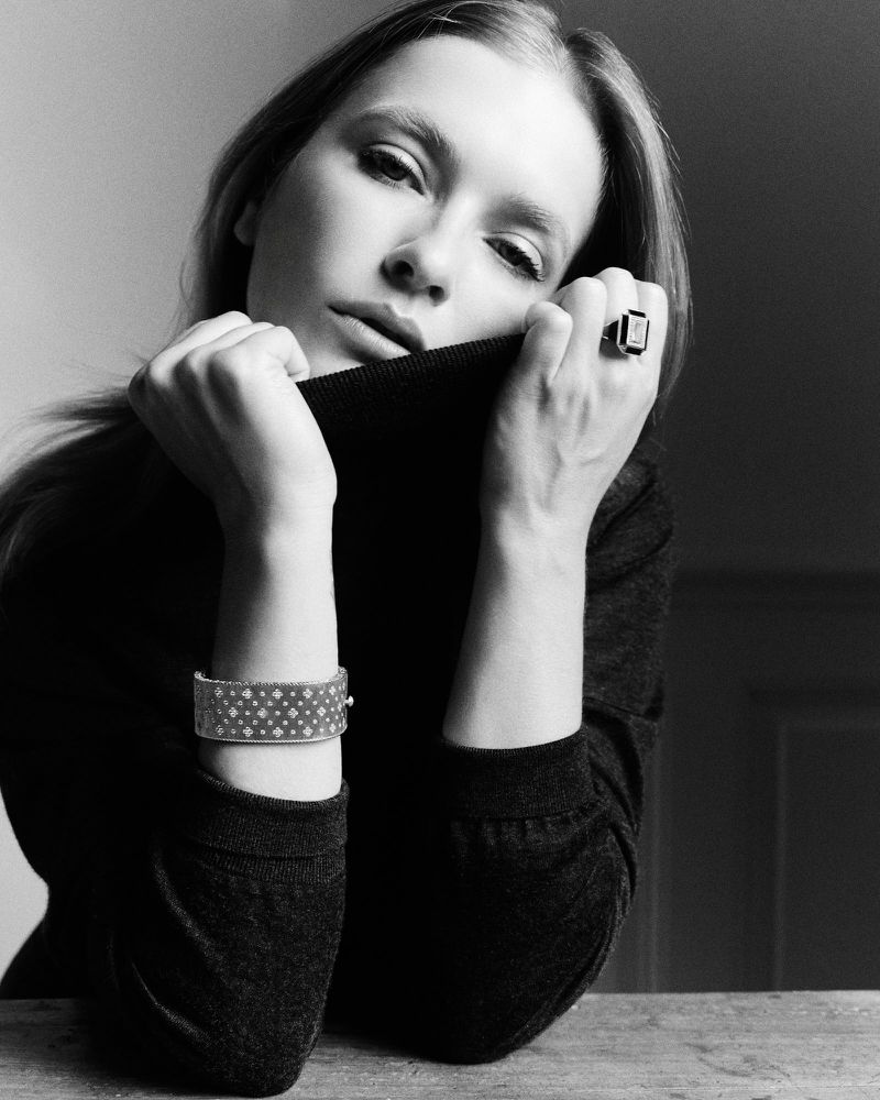 Monochrome portrait of a contemplative woman wearing jewelry, with a dark top, fair skin, and neutral studio background, shot at eye-level.