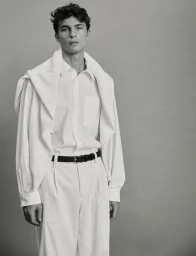 Model Yulef Bopp wearing a white shirt and trousers with a sweater draped over his shoulders against a grey studio backdrop.