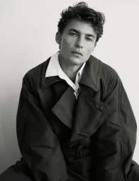 Model Yulef Bopp wearing a dark coat and white shirt against a neutral background in a black-and-white editorial portrait.