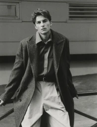 Man wearing a grey wool overcoat and collared shirt leaning against an outdoor railing in a black and white editorial portrait.