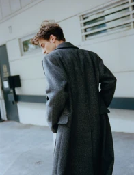 Model Yulef Bopp wearing a dark grey wool coat outdoors against an urban building exterior background.