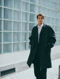 Model Yulef Bopp wearing a textured dark coat against a glass block wall for The Modern Suit editorial project
