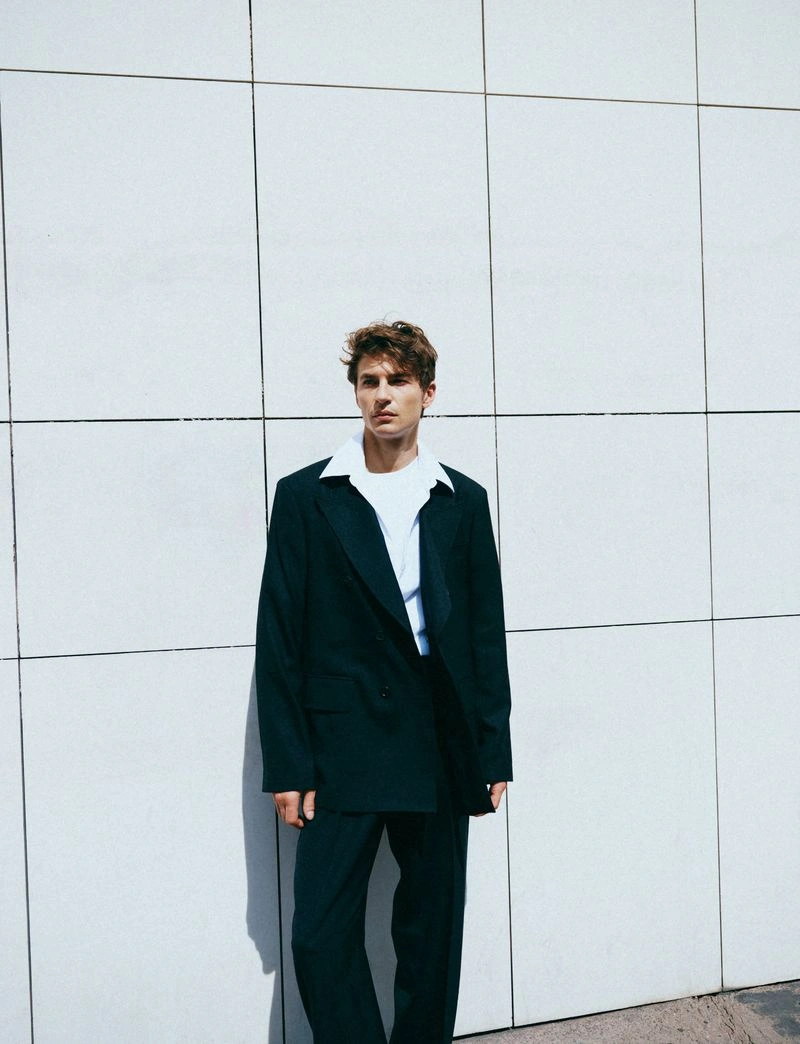 Model wearing a dark oversized suit and white shirt standing against a tiled wall