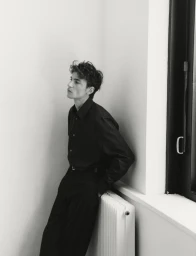 Model Yulef Bopp wearing a dark shirt and trousers leaning against a white wall next to a window.