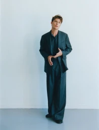 Model Yulef Bopp wearing a dark grey suit and shirt standing against a light background for The Modern Suit editorial project.