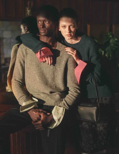Man in a grey sweater and woman in a dark green sweater posing together for a fashion editorial shot.