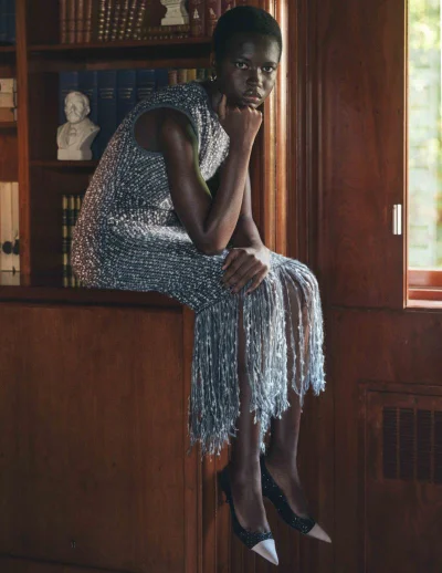 Model seated on a wooden bookshelf wearing a grey sleeveless embellished dress and black heels for L'Officiel Italia.