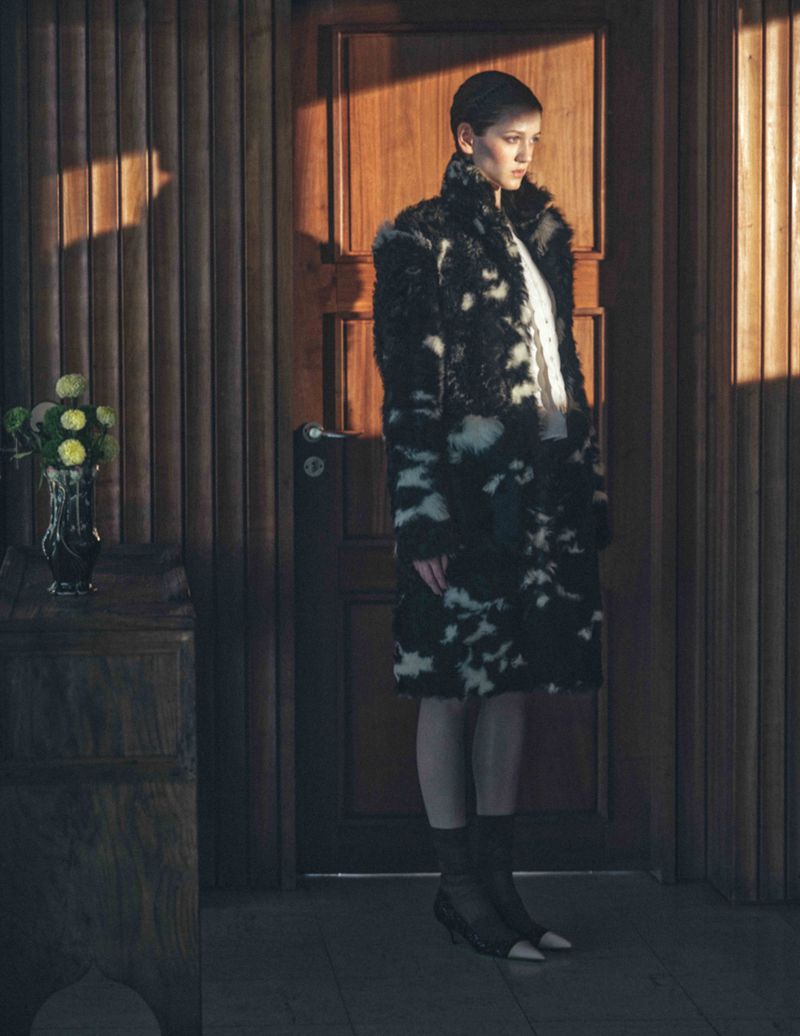 Model wearing a black and white patterned faux fur coat standing near a wooden door in a warmly lit interior setting.