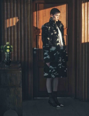 Model wearing a black and white patterned faux fur coat standing near a wooden door in a warmly lit interior setting.