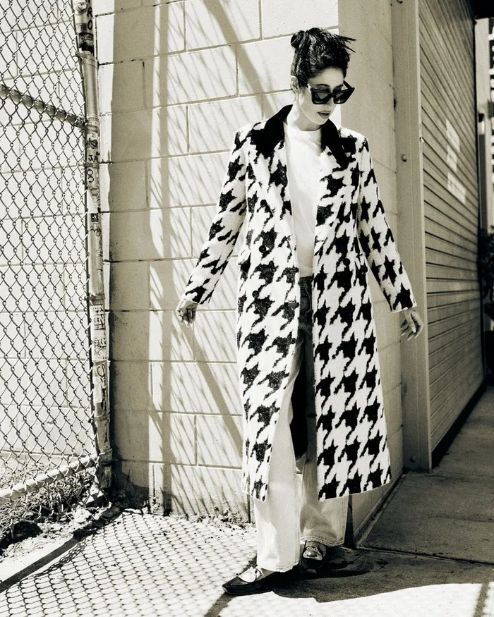 Model in a black and white houndstooth coat and sunglasses standing against a brick wall near a chain-link fence.