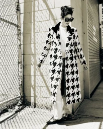 Model in a black and white houndstooth coat and sunglasses standing against a brick wall near a chain-link fence.