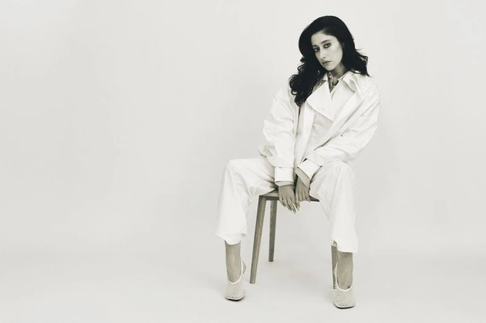 Woman with long dark hair wearing a white outfit seated on a wooden chair against a plain light background