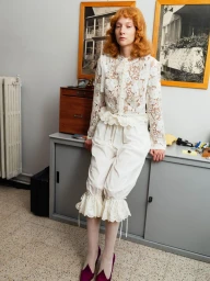 Model with red hair leaning against a grey cabinet wearing a white lace blouse and ruffled trousers