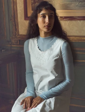 Model with long dark wavy hair seated indoors wearing a light blue top and white sleeveless dress with embroidery.