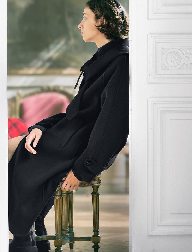 Woman in a black coat seated on an ornate stool framed by a white doorway for ICICLE Autumn/Winter 2026