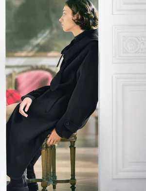 Woman in a black coat seated on an ornate stool framed by a white doorway for ICICLE Autumn/Winter 2026
