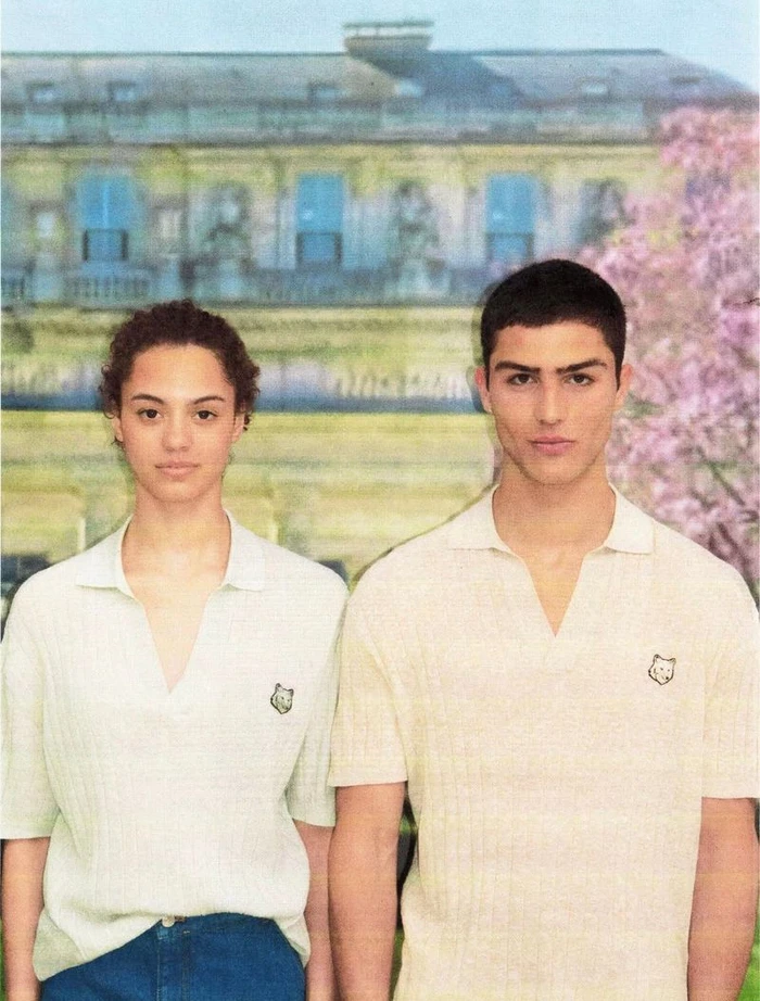 Two models wearing white polo shirts standing in front of a printed backdrop of a building and trees.