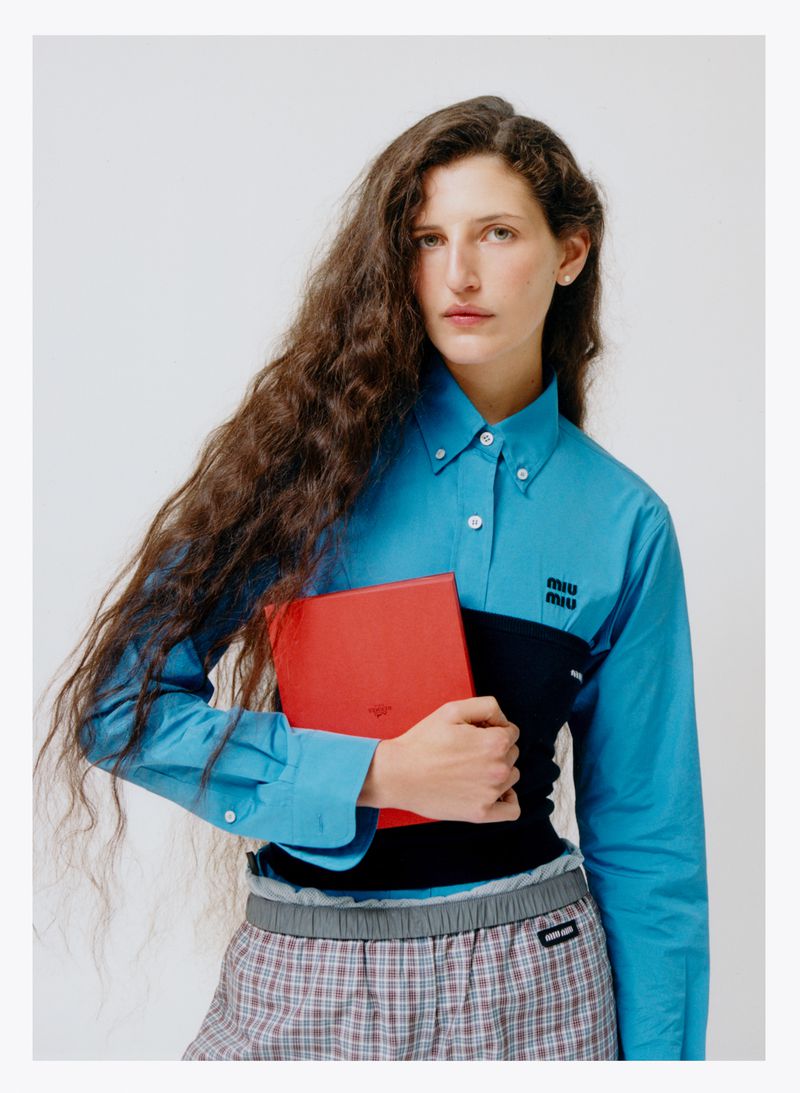 A female fashion model poses indoors, wearing a Miu Miu turquoise button-down shirt, plaid skirt, and holding a red book against a white backdrop.