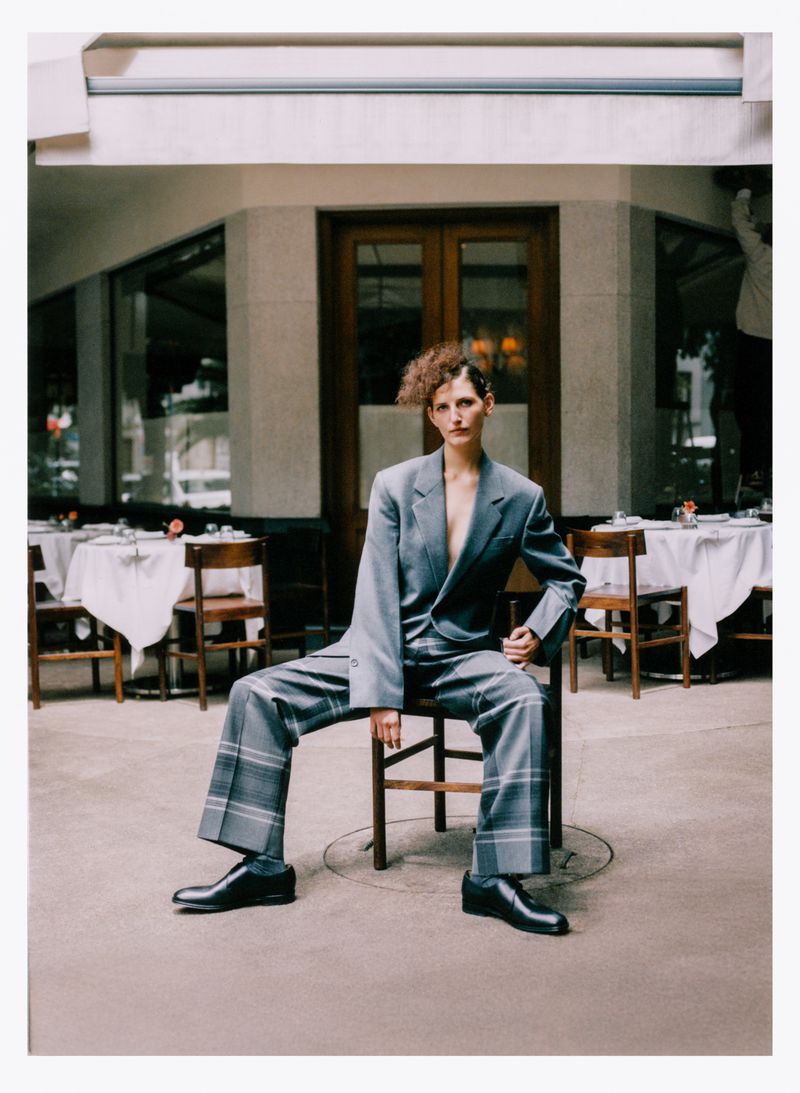 Fashionable woman in a grey pantsuit poses sitting on a chair outside a restaurant with tables and chairs on the patio.