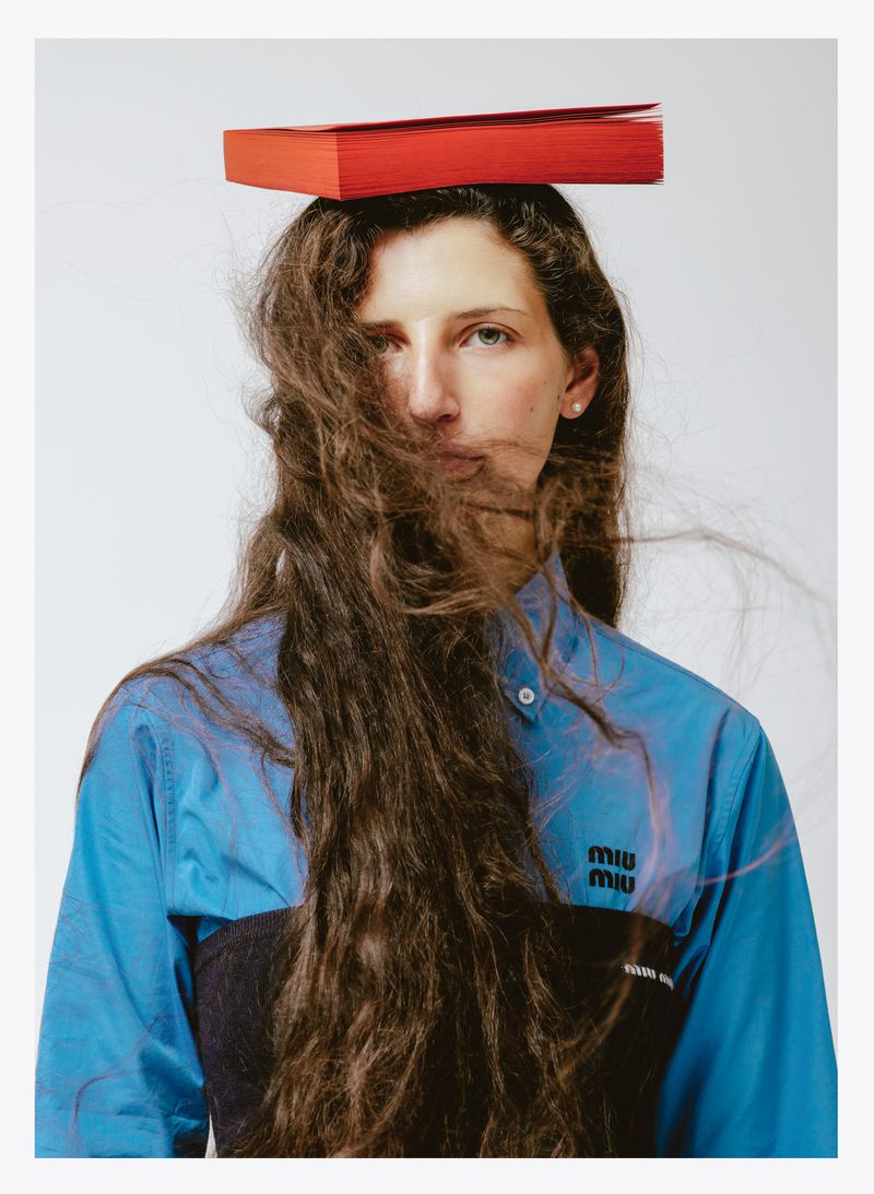 Young woman with long brown hair wearing a blue collared shirt with a red book on her head in a studio setting with white background.