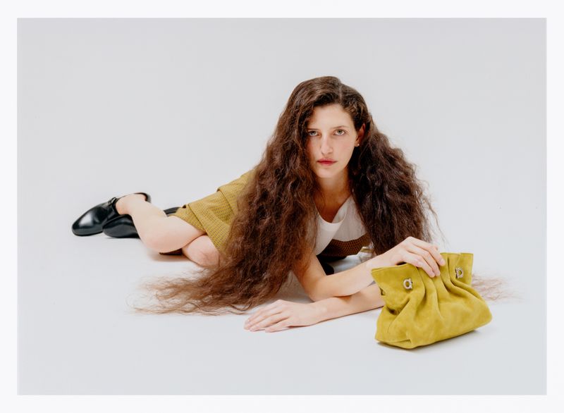 A woman with long curly hair poses with a yellow purse on a white background, wearing a tan shirt and black shoes.