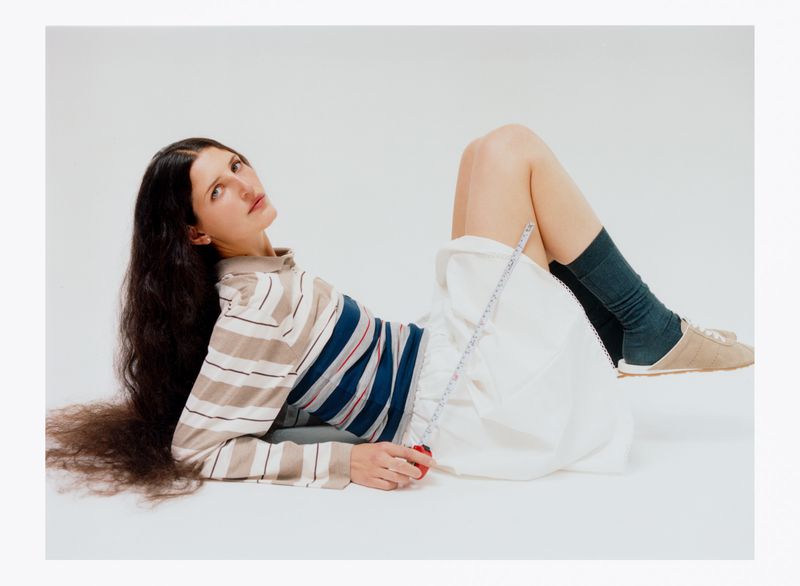 An adult female is reclining on a white surface, wearing a striped top and white skirt, long hair spread around her.