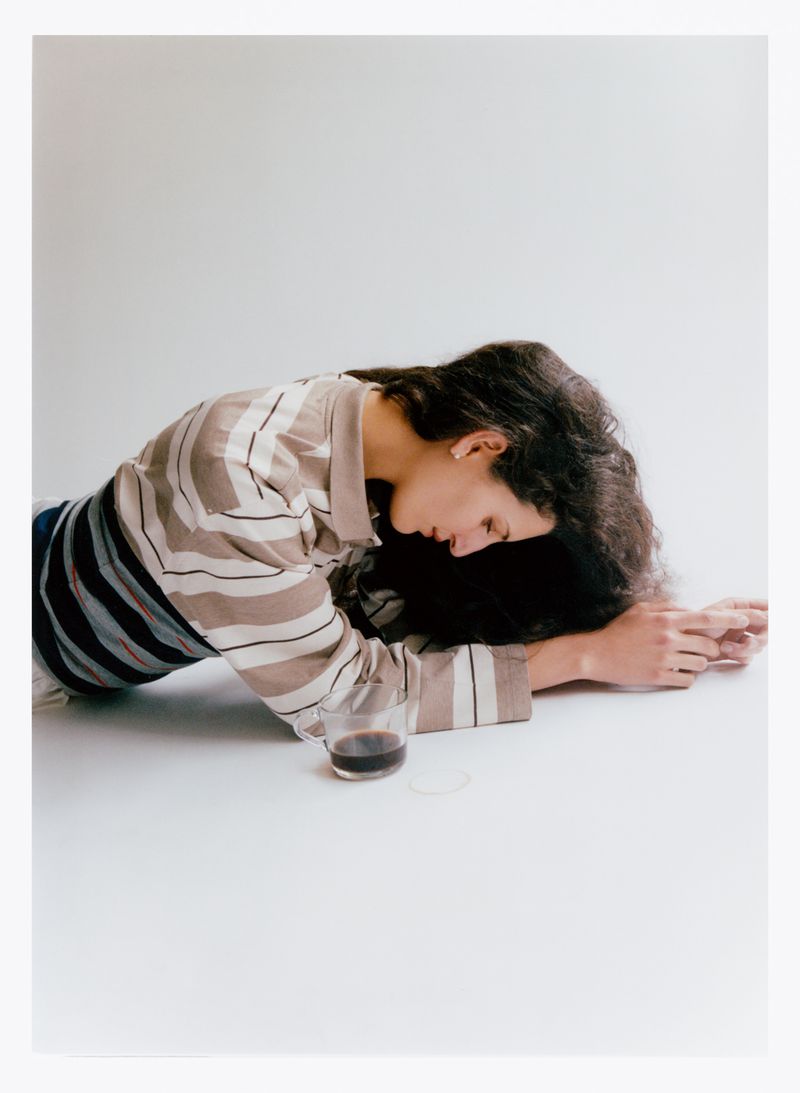 Brunette woman lying on a white surface with a glass of dark liquid. She wears a striped shirt in a minimalist, indoor setting.