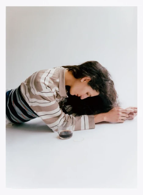Brunette woman lying on a white surface with a glass of dark liquid. She wears a striped shirt in a minimalist, indoor setting.