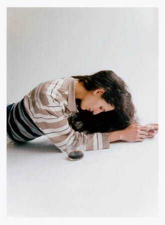 Brunette woman lying on a white surface with a glass of dark liquid. She wears a striped shirt in a minimalist, indoor setting.
