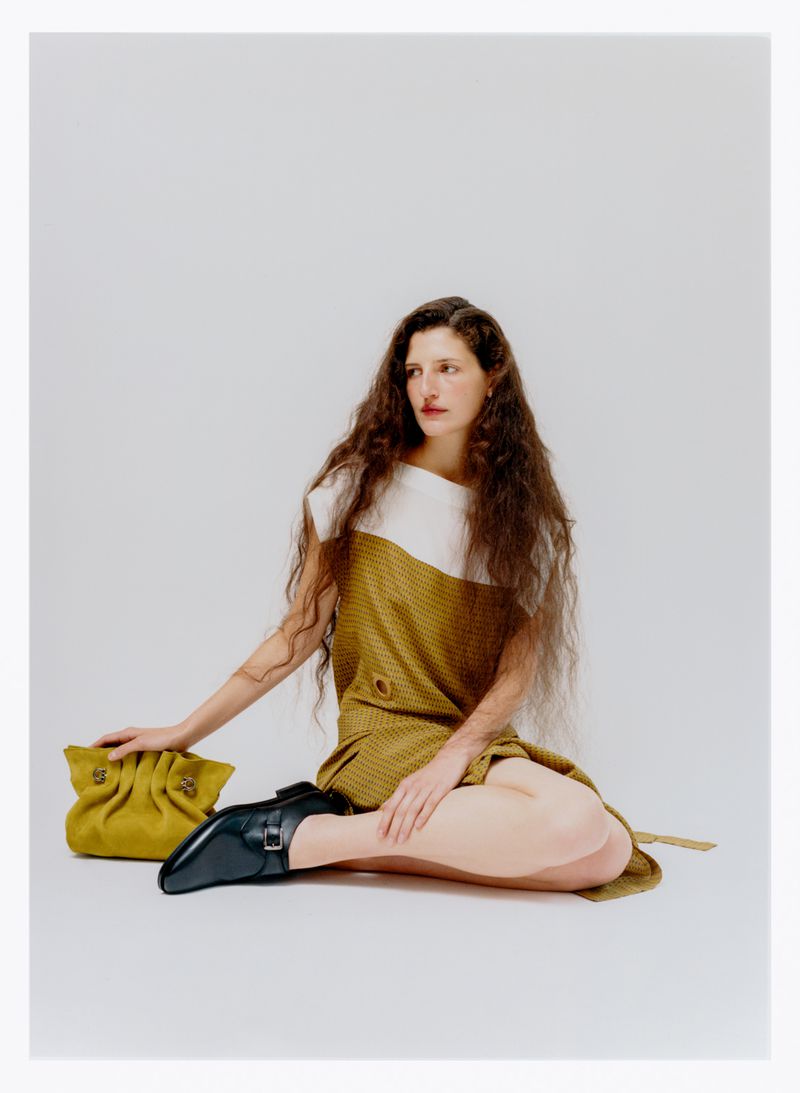 A woman with long curly hair poses in a brown and white dress with a yellow purse and black shoe against a white backdrop indoors.