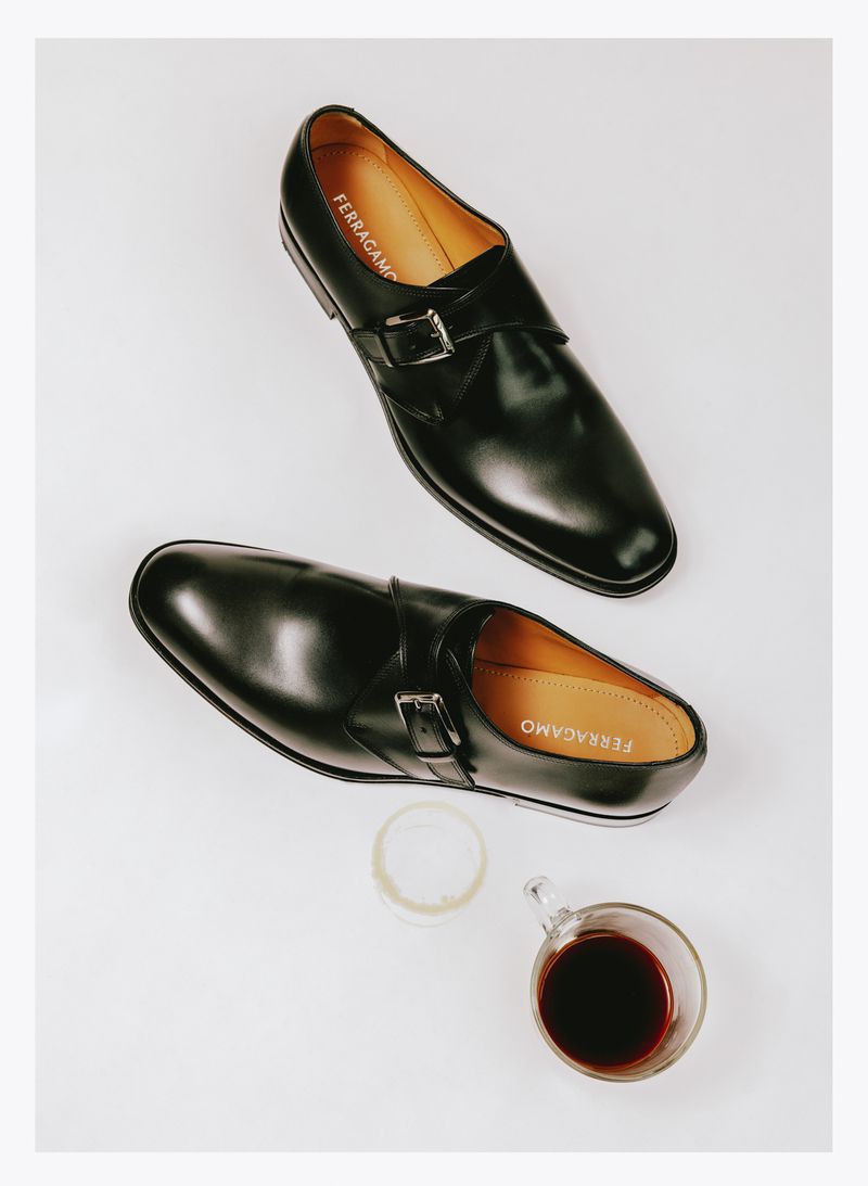 Elevated view of two black leather Ferragamo Monk Strap shoes with silver buckles alongside a glass of dark liquid on a white surface.