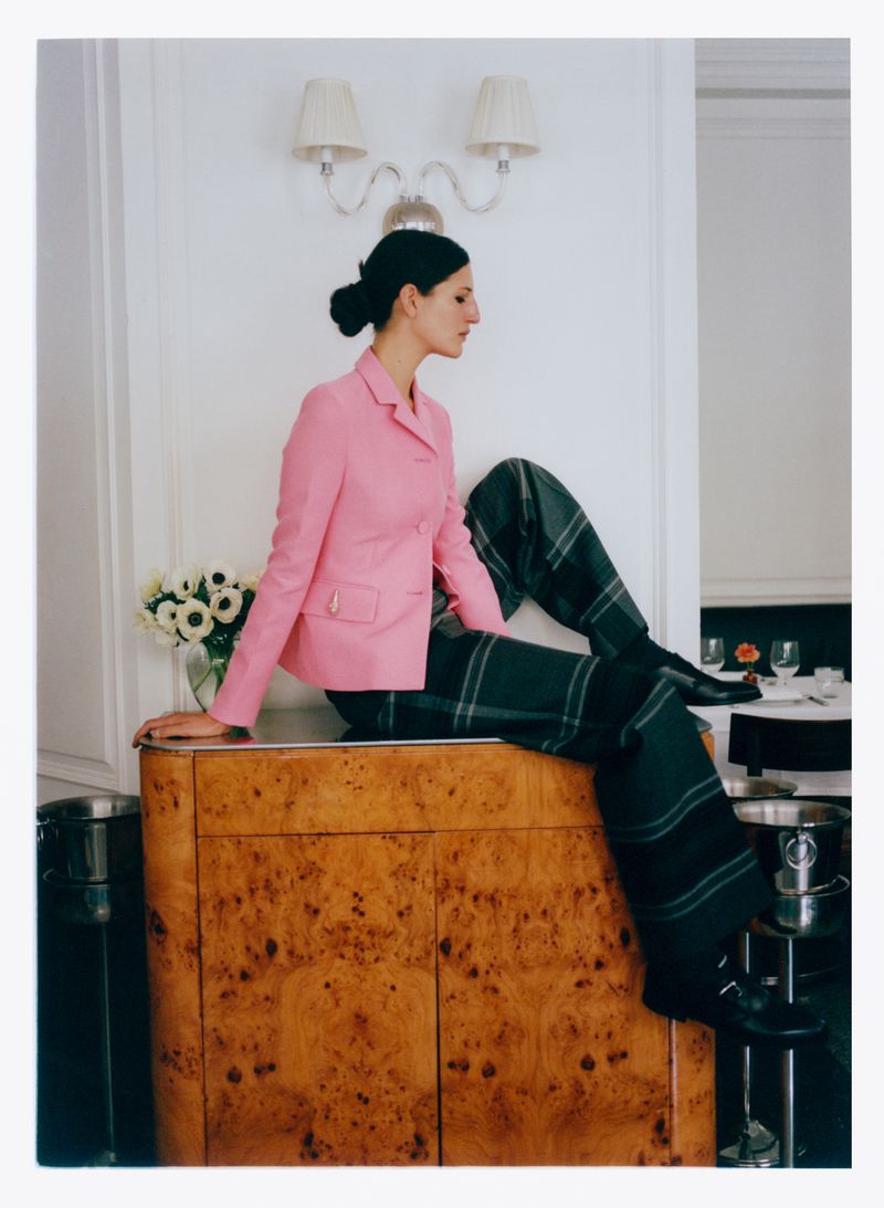Woman in pink blazer and plaid pants sits on a wooden cabinet with white flowers in a vase, against a white wall with sconces.