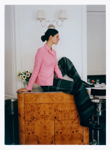 Woman in pink blazer and plaid pants sits on a wooden cabinet with white flowers in a vase, against a white wall with sconces.