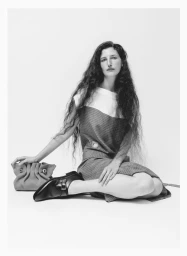 Woman with long dark hair seated on a white floor holding a handbag wearing a structured dress and black loafers