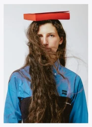 Woman with long wavy hair balancing a red book on her head while wearing a blue shirt against a white backdrop.