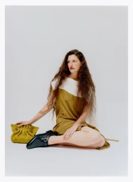 Woman with long curly brown hair seated wearing a two-tone dress holding an olive pouch against a plain white background