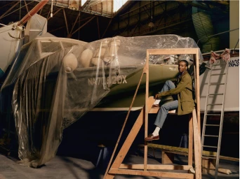 Black woman seated on a wooden frame structure in a boathouse beside a boat covered in plastic sheeting
