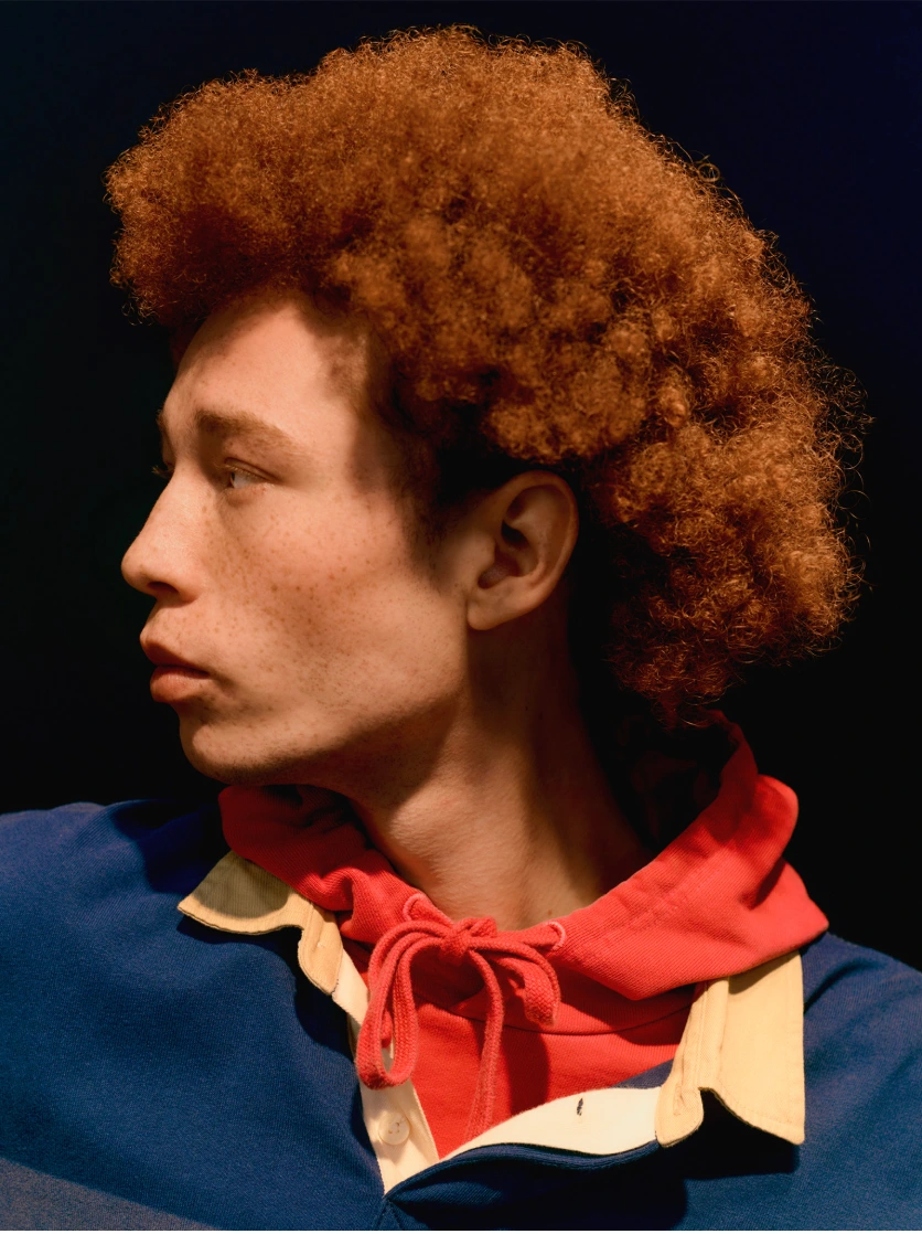 Profile portrait of a young man with curly red hair and freckles wearing a blue and red polo shirt against a dark background.