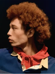 Profile portrait of a young man with curly red hair and freckles wearing a blue and red polo shirt against a dark background.