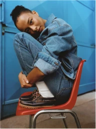Model wearing a denim jacket and jeans while crouched on a bright red chair against a blue wall
