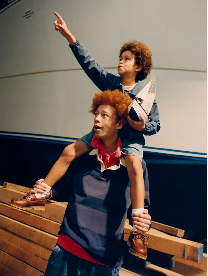 Young person carrying a child on their shoulders holding a model boat in front of a large white hull and wooden planks